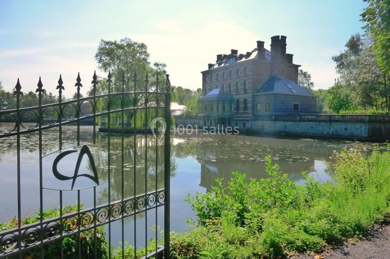 Manoir en pierre au bord d'un étang, entouré de végétation, avec une grille en fer forgé au premier plan.
