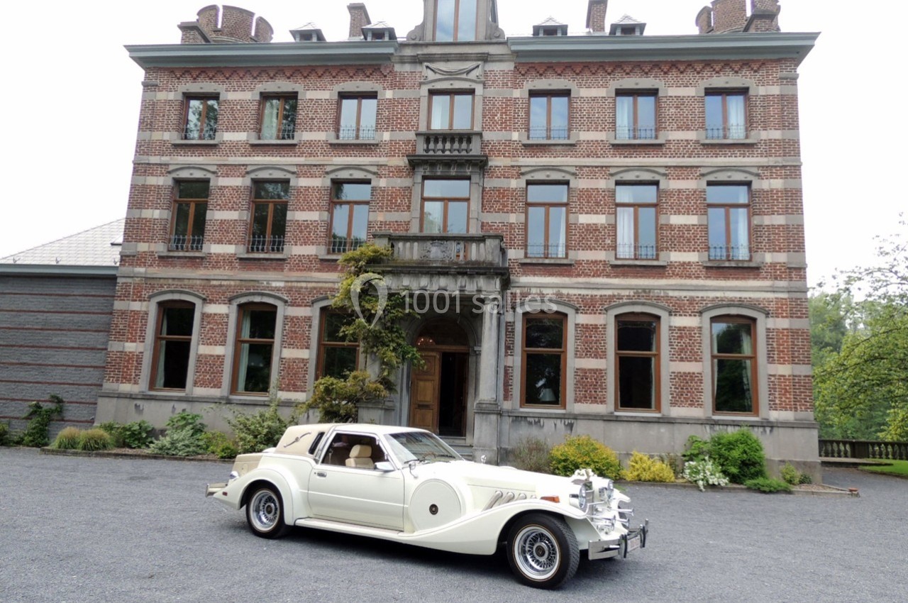 Voiture ancienne blanche garée devant une grande maison en briques rouges avec des fenêtres symétriques.