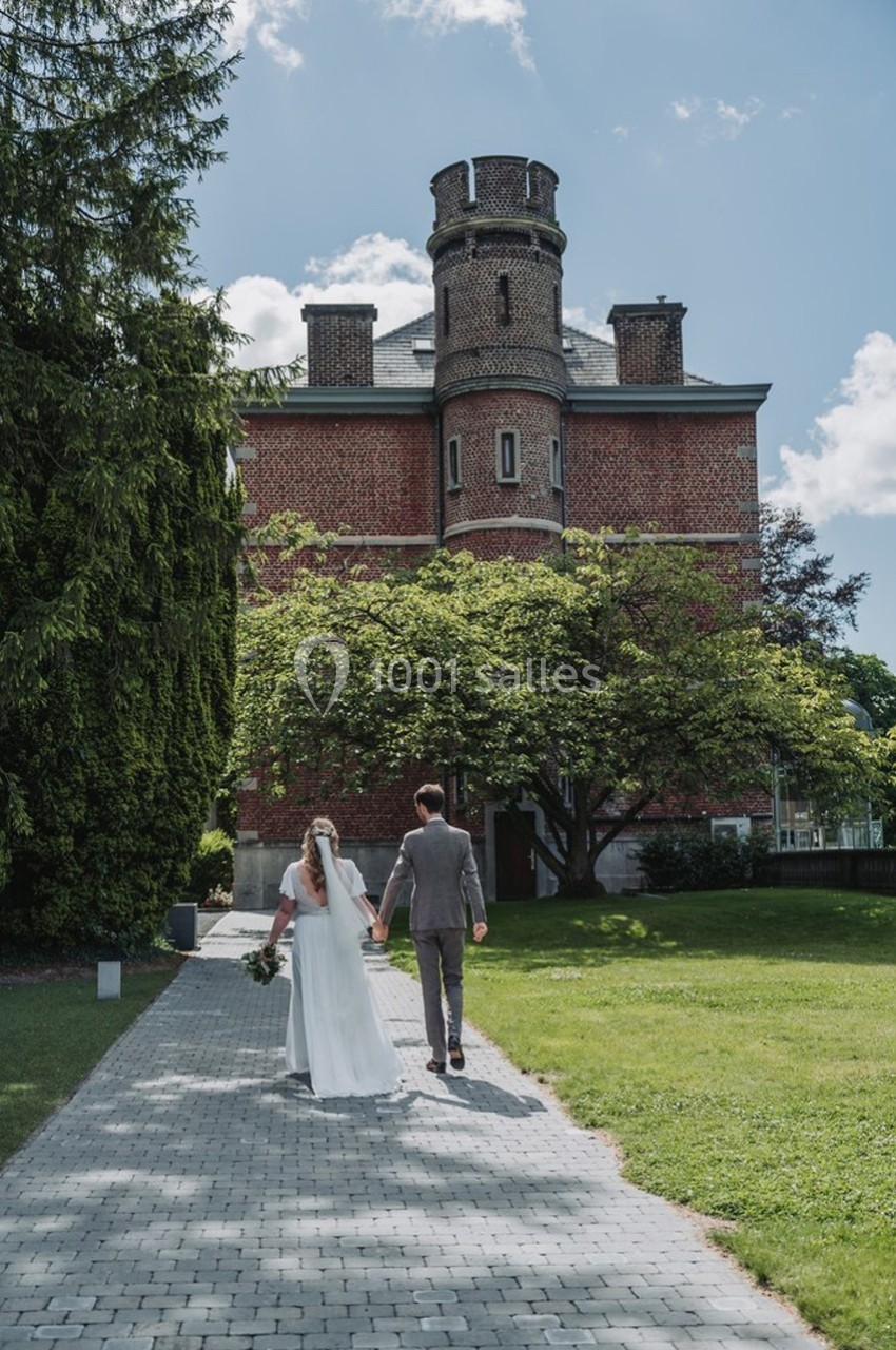 Un couple en tenue de mariage marche sur une allée pavée devant un bâtiment en briques entouré de verdure.