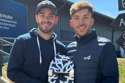 Deux hommes souriants posent à l'extérieur, l'un tenant un skateboard noir et blanc devant un bâtiment moderne.