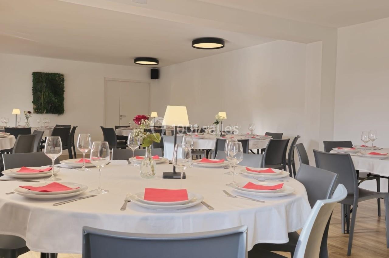 Salle de restaurant lumineuse avec tables rondes dressées, nappes blanches, serviettes rouges et décoration sobre.