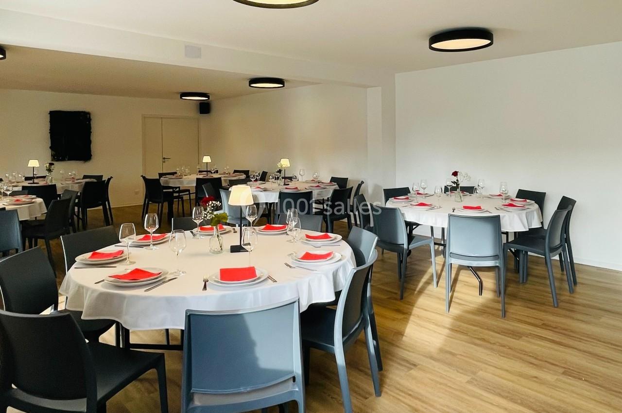 Salle de réception avec tables rondes dressées, nappes blanches, serviettes rouges et chaises grises, éclairage moderne.