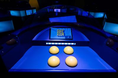 Poste de jeu interactif avec quatre boutons jaunes et un écran affichant ’Quiz Room’ dans un éclairage bleu.