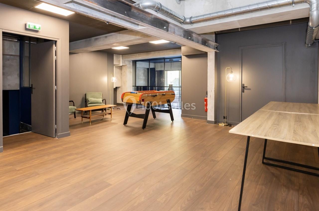 Espace intérieur moderne avec parquet, baby-foot, fauteuils et éclairage industriel.