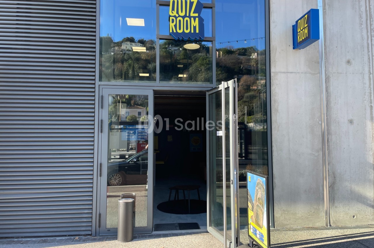 Entrée d'un bâtiment avec une porte vitrée ouverte, une enseigne ’Quiz Room’ et un panneau publicitaire devant.