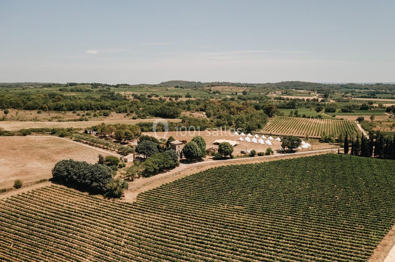 Vue aérienne d'un paysage rural avec des vignobles, des arbres, des bâtiments et des tentes blanches sous un ciel dégagé.