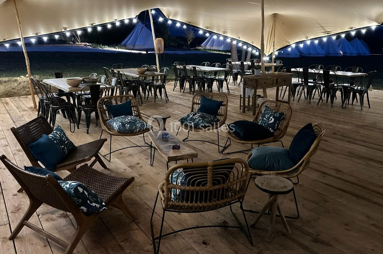 Espace extérieur couvert avec fauteuils en rotin, tables et chaises, éclairé par des guirlandes lumineuses la nuit.