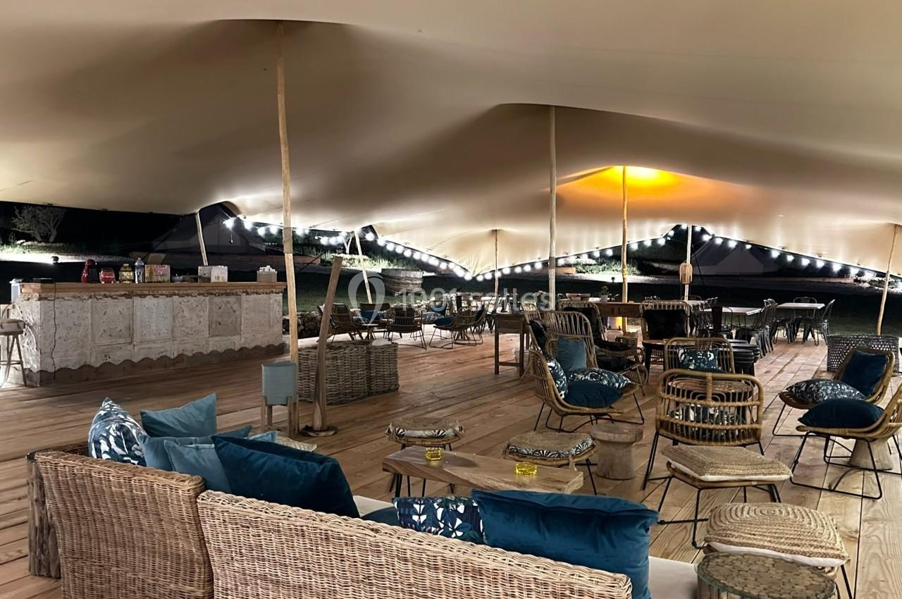 Espace extérieur couvert avec mobilier en osier, tables basses et éclairage chaleureux sous une tente tendue.