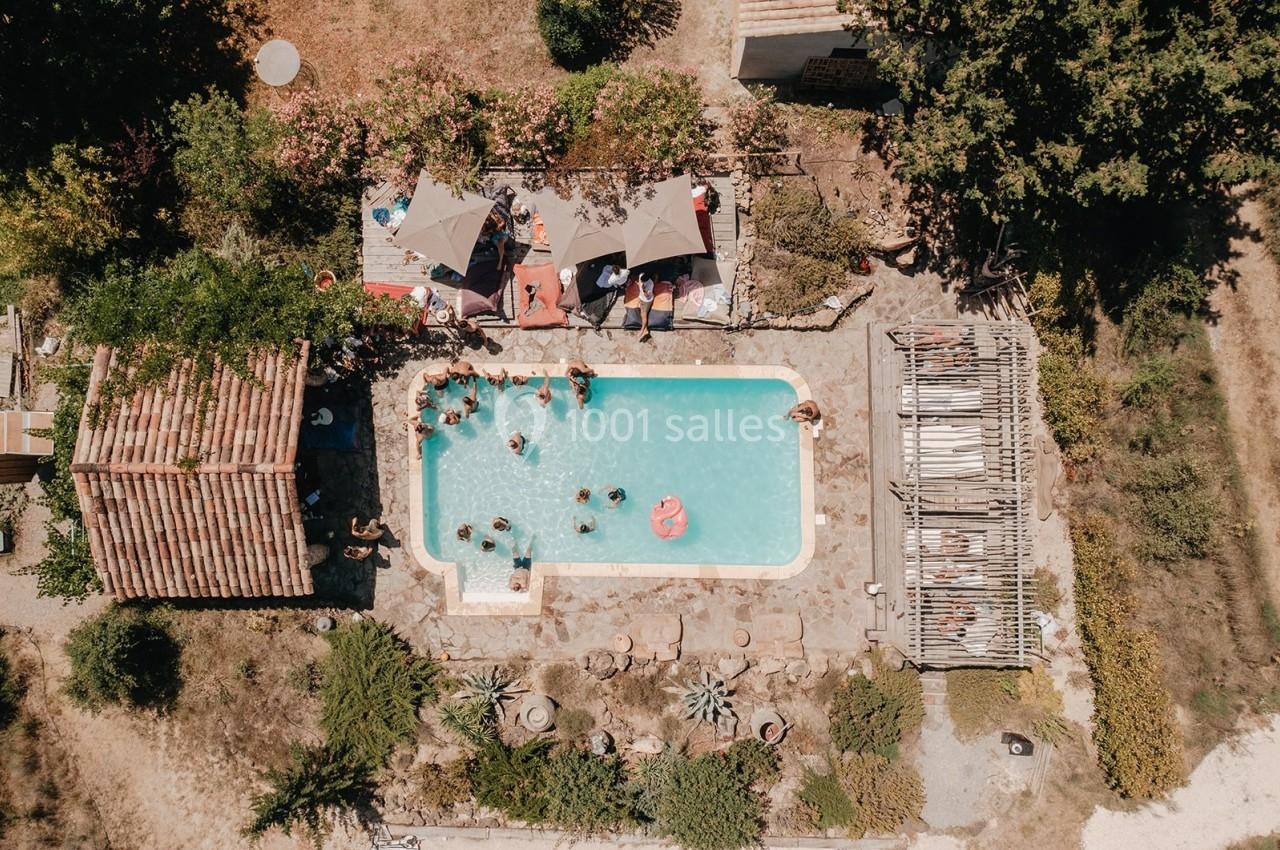 Vue aérienne d'une piscine entourée de végétation, avec des personnes se baignant et des parasols à proximité.