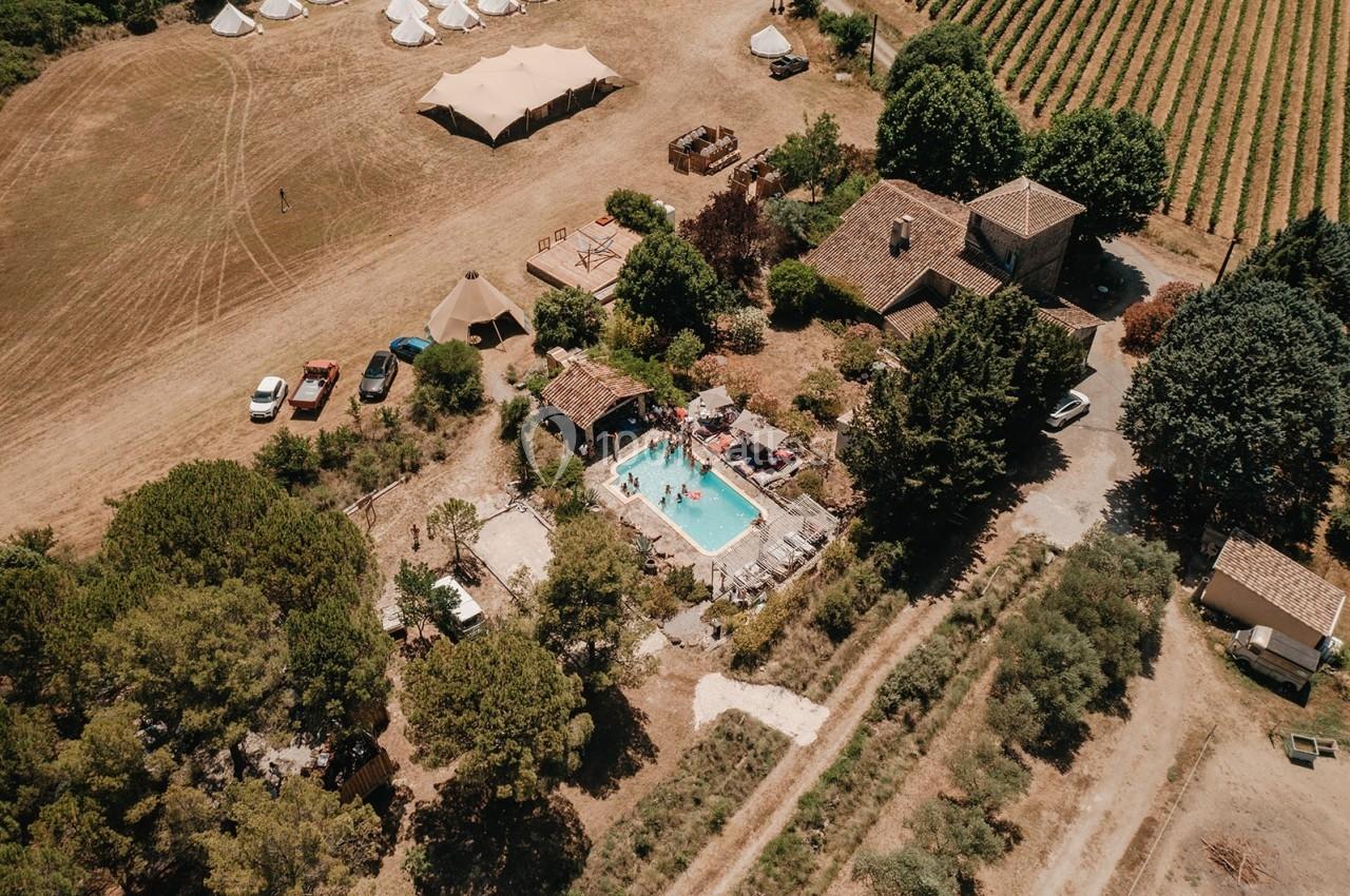 Vue aérienne d'une propriété rurale avec piscine, entourée de vignes, arbres et tentes dans un paysage ensoleillé.