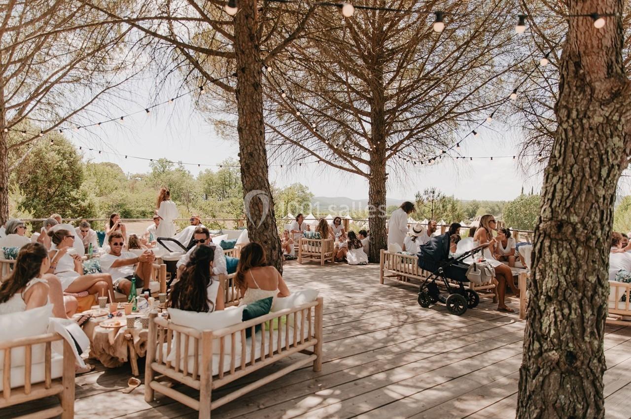 Des personnes assises sur des bancs en bois sous des arbres, dans un espace extérieur décoré de guirlandes lumineuses.