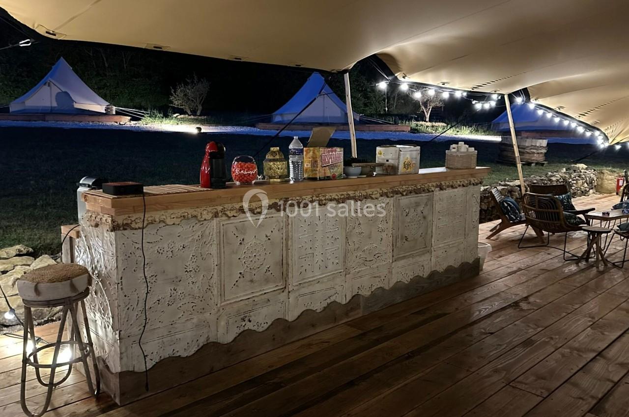 Comptoir en bois décoré sous une tente éclairée, avec des boissons et des collations, entouré de tables et de chaises.