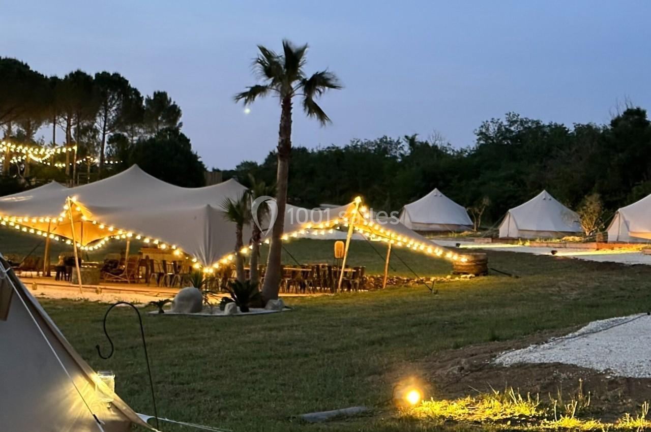 Tentes éclairées par des guirlandes lumineuses dans un espace extérieur en soirée, entouré de végétation.