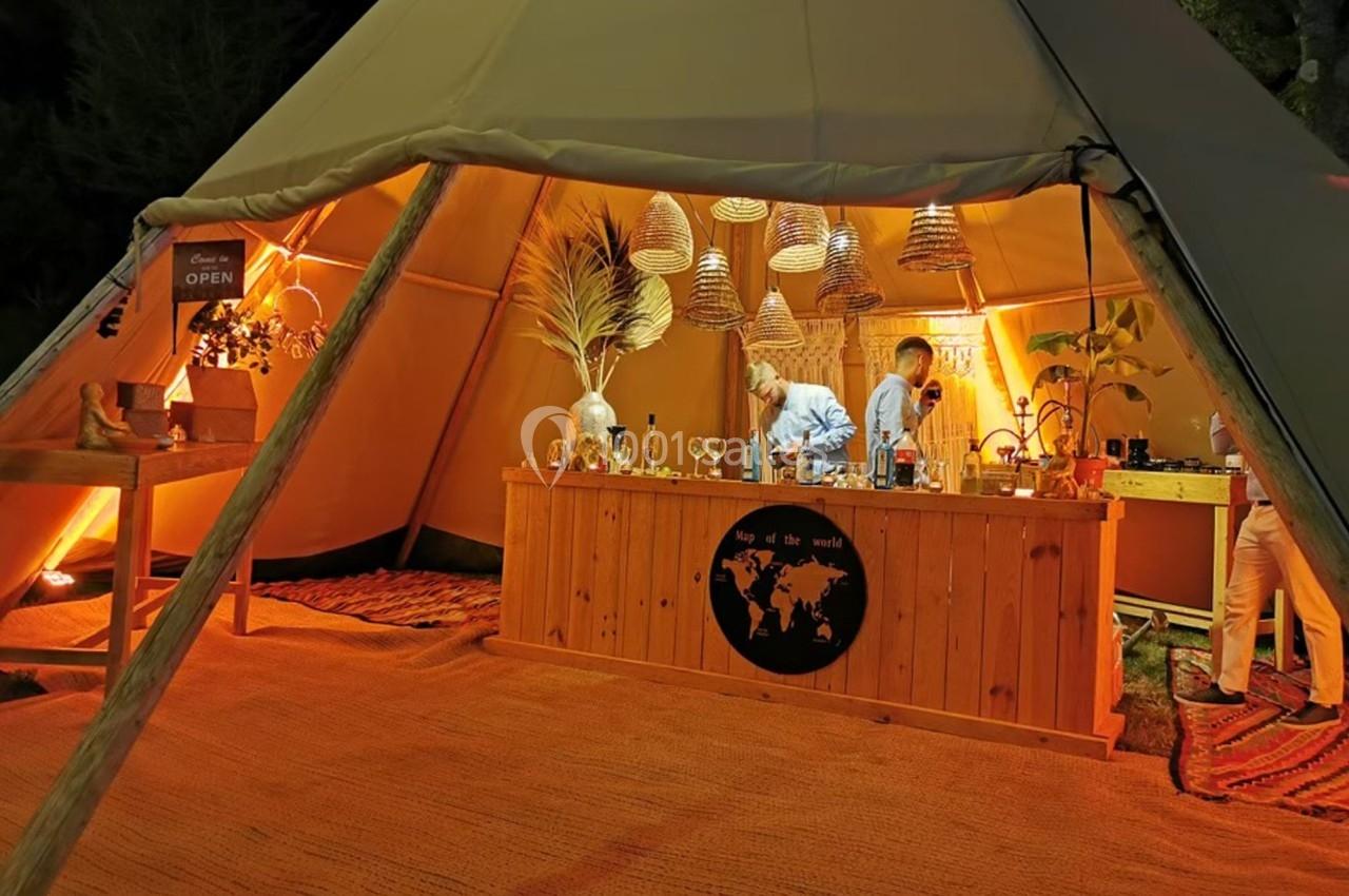 Un bar en bois éclairé à l'intérieur d'une tente, avec deux personnes préparant des boissons sous des lampes suspendues.