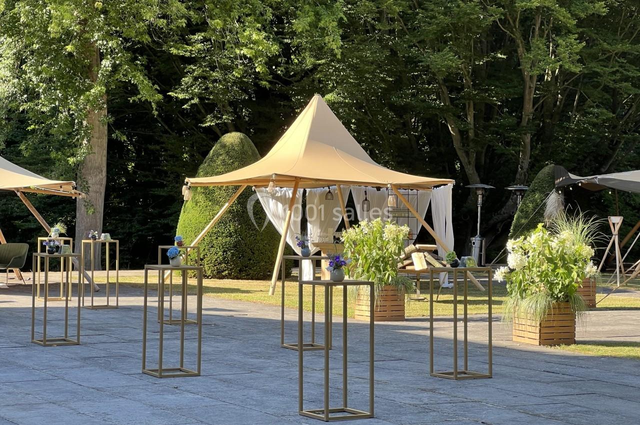 Tente beige élégante installée dans un jardin verdoyant, entourée de tables hautes décorées de fleurs.
