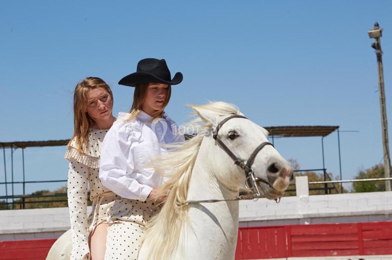 Deux jeunes femmes montent un cheval blanc dans une arène en plein air, sous un ciel dégagé.