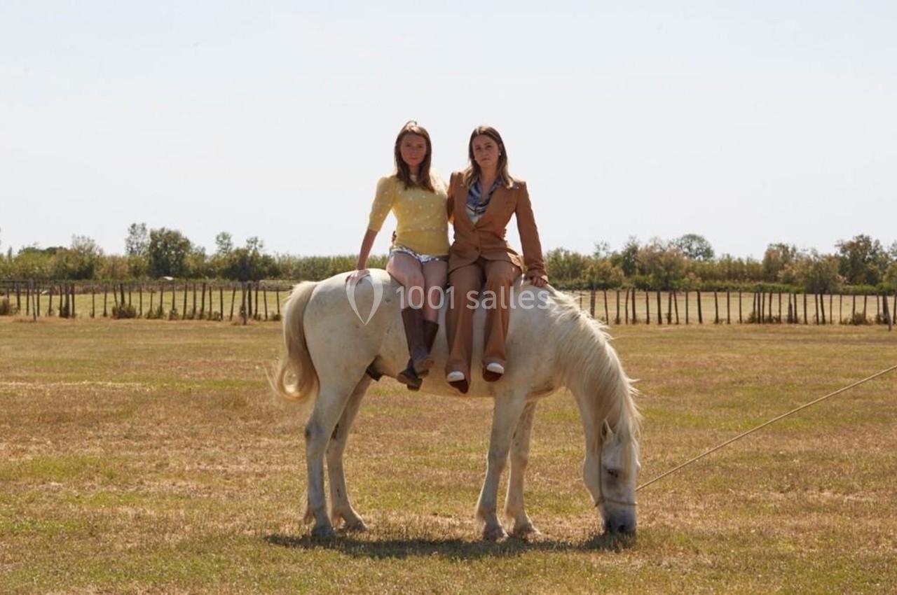 Deux femmes assises côte à côte sur un cheval blanc broutant dans un champ dégagé par temps ensoleillé.