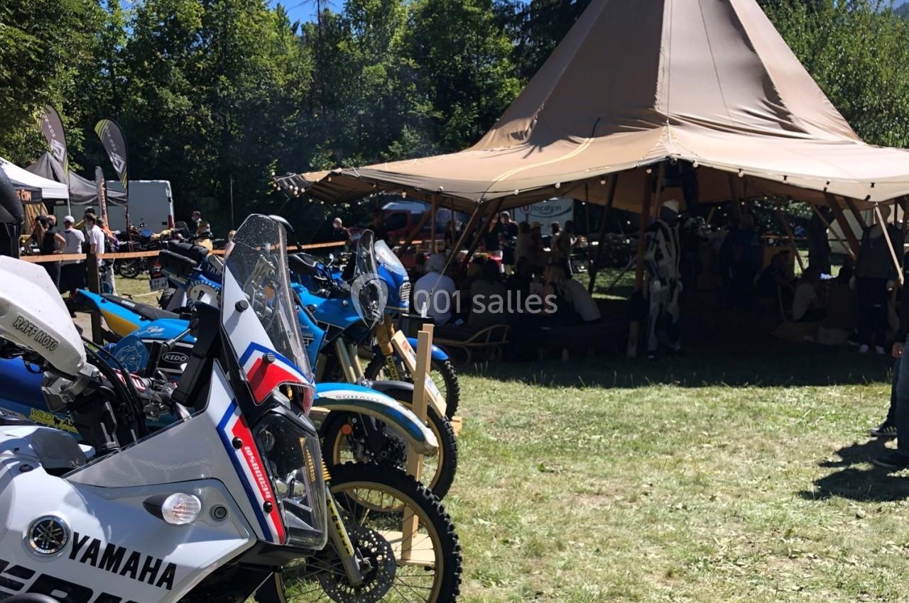 Plusieurs motos garées sur une pelouse près d'une grande tente beige, avec des personnes rassemblées à l'ombre.