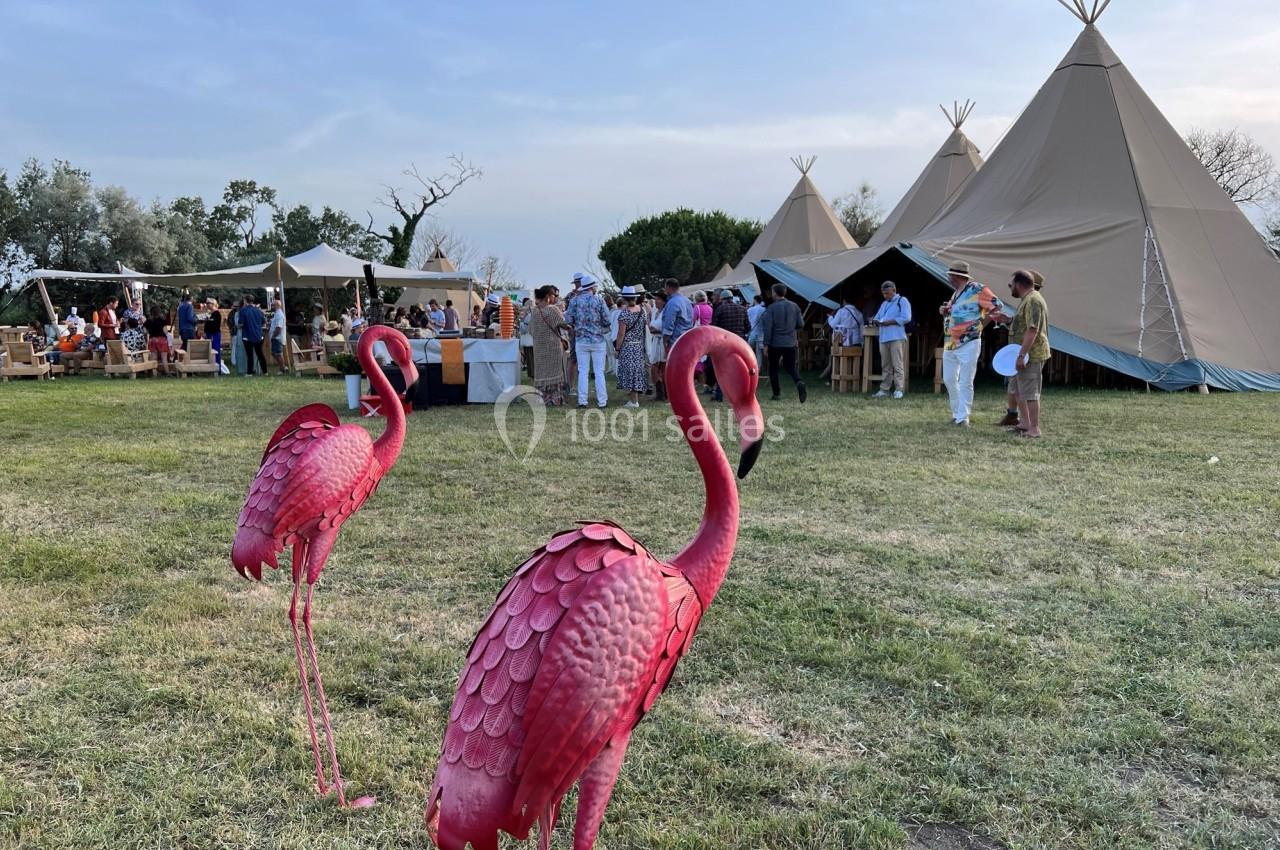 Deux statues de flamants roses en métal sur une pelouse, avec des tipis et des personnes en arrière-plan.