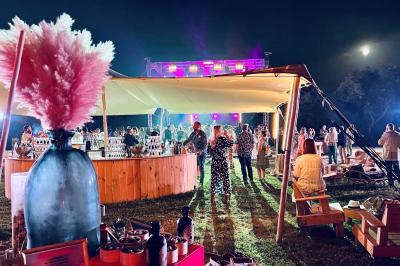 Fête nocturne en plein air avec un bar central, des invités debout et des lumières colorées sous une tente éclairée.