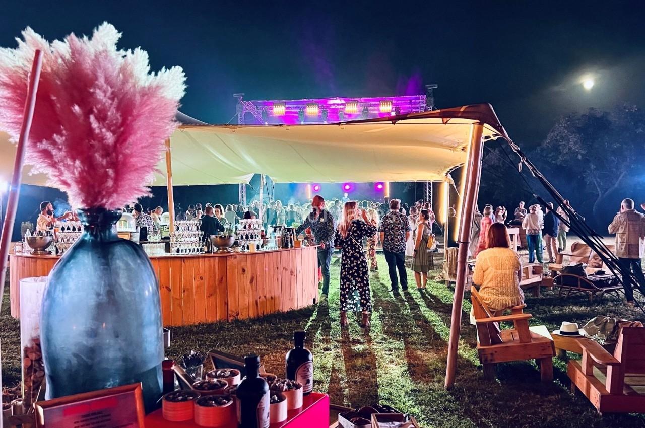 Fête nocturne en plein air avec un bar central, des invités debout et des lumières colorées sous une tente éclairée.