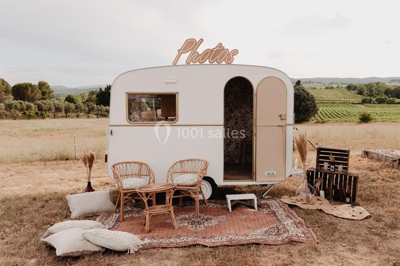 Caravane aménagée en cabine photo, entourée de mobilier en rotin et tapis, située dans un champ ensoleillé.