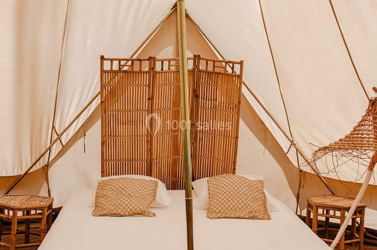 Lit double avec coussins beiges dans une tente équipée de cloisons en bambou et de petites tables en bois.