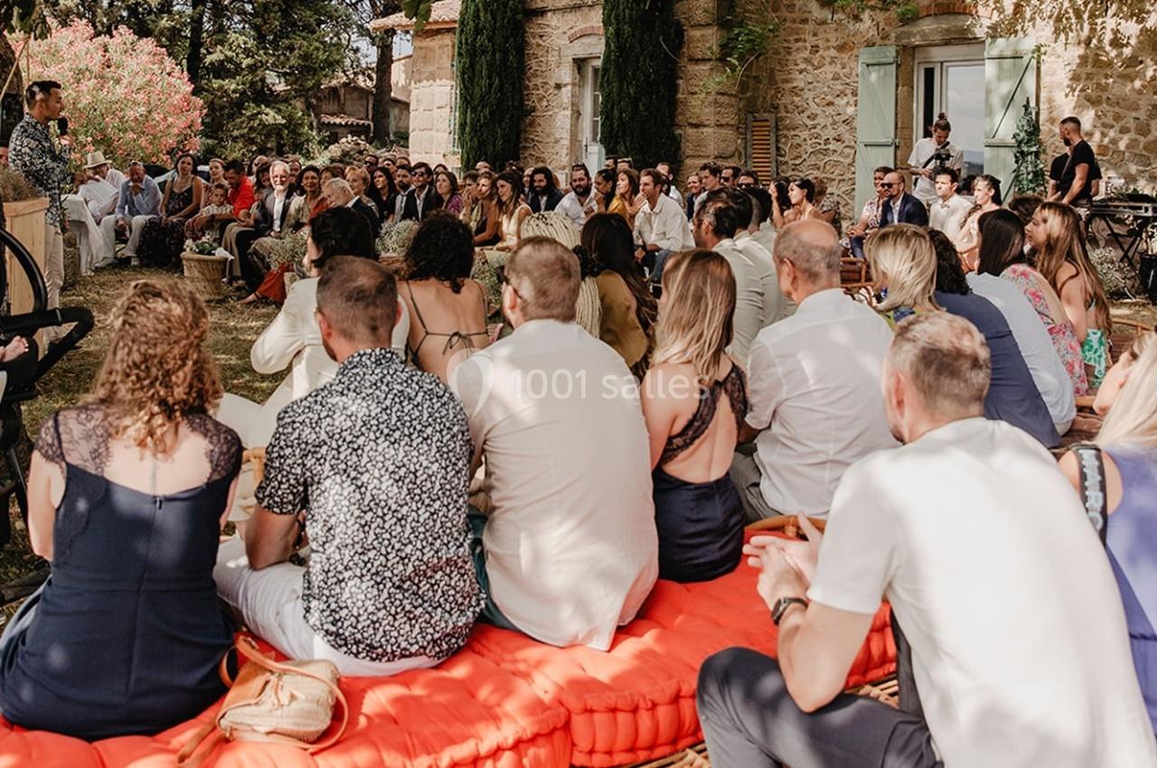 Un groupe de personnes assises en plein air, écoutant un orateur devant une maison en pierre entourée de végétation.