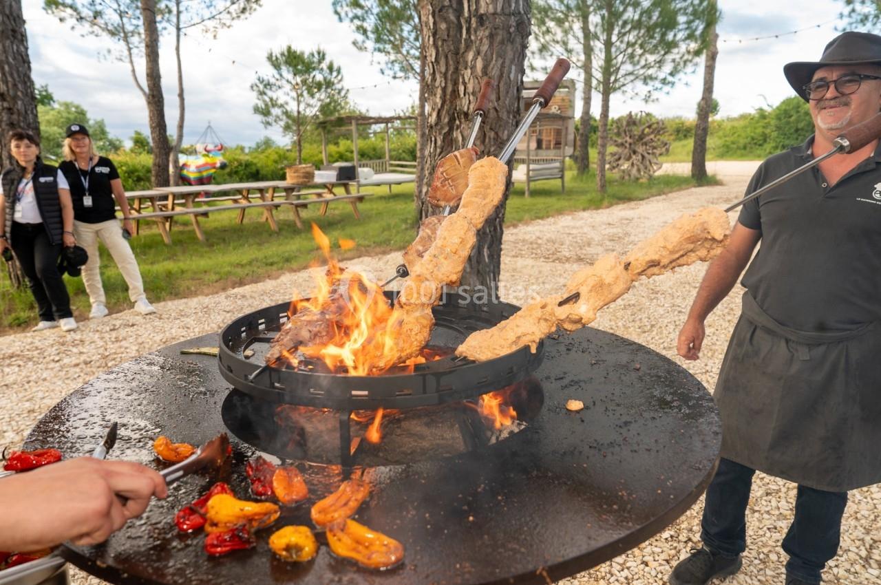 Des brochettes de viande et des légumes grillent sur un feu en plein air, entourés de convives et d'arbres.