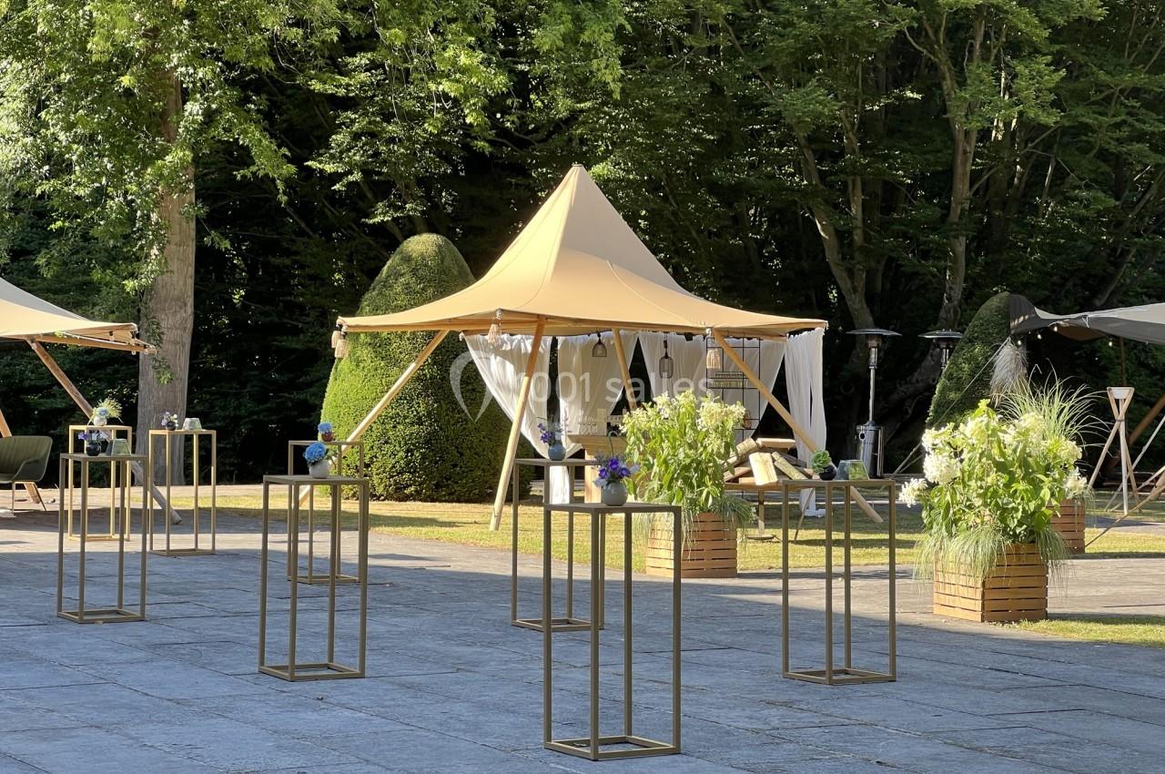 Tentes élégantes installées dans un jardin avec des tables hautes décorées de fleurs et une végétation en arrière-plan.