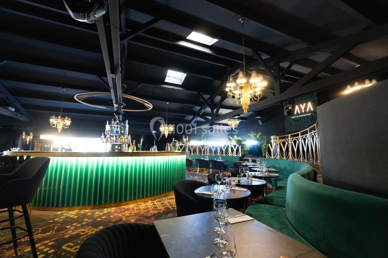 Intérieur d'un bar élégant avec comptoir lumineux vert, tables dressées et décorations modernes sous un plafond industriel.