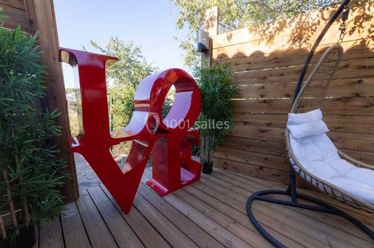 Sculpture rouge ’LOVE’ sur une terrasse en bois entourée de plantes et d'un fauteuil suspendu.