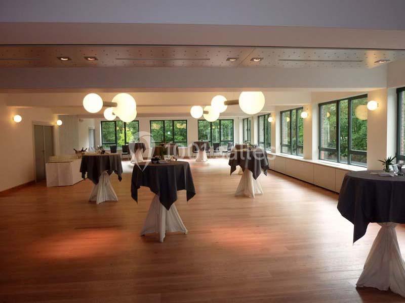 Salle lumineuse avec parquet, grandes baies vitrées, tables hautes nappées de gris et éclairage suspendu moderne.