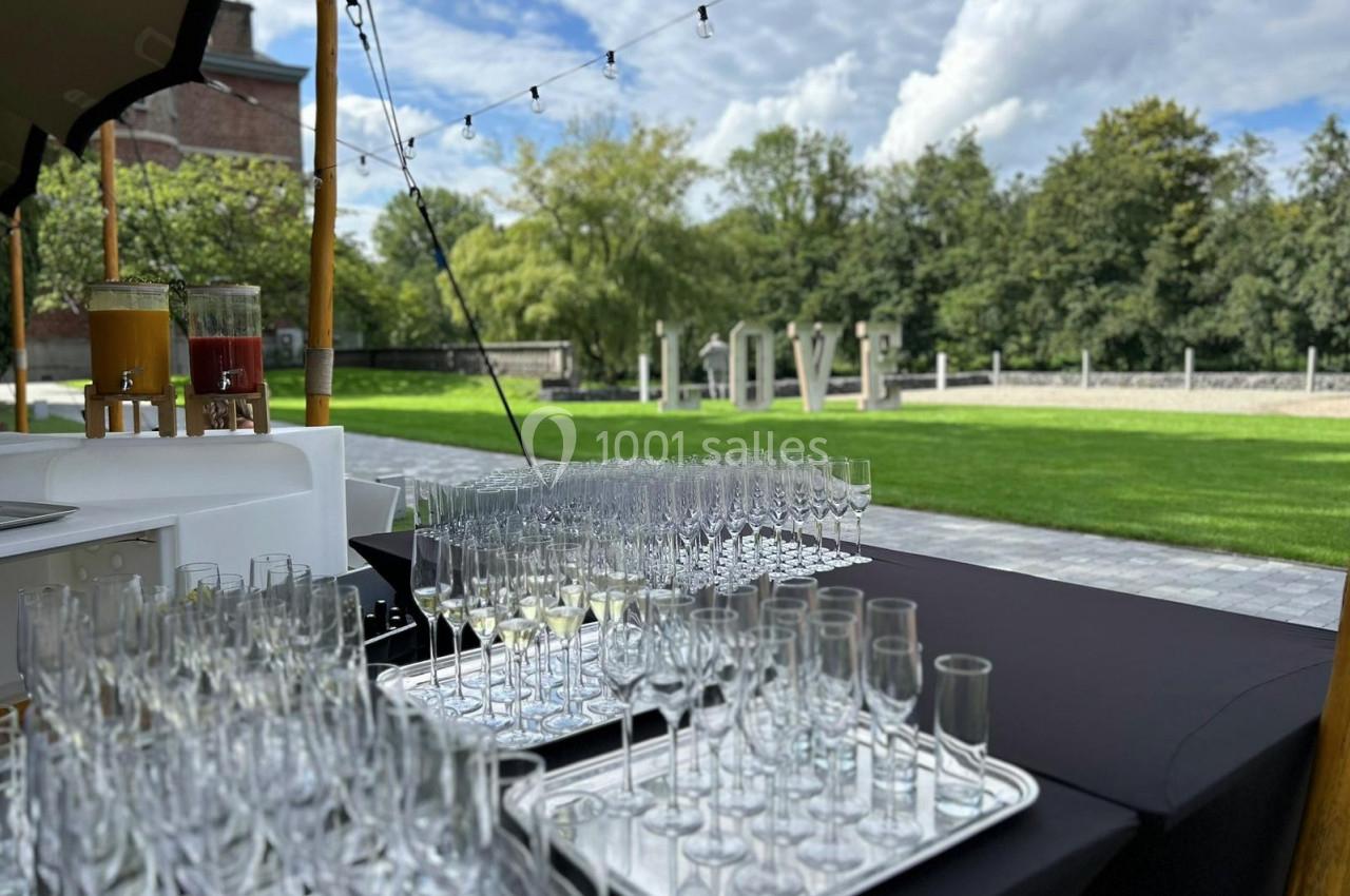 Verres à pied alignés sur des plateaux sur une table noire en extérieur, avec un jardin et un ciel nuageux en arrière-plan.