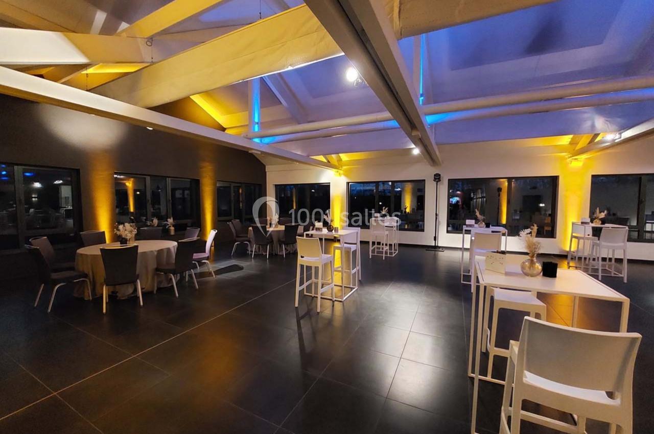 Salle spacieuse avec tables et chaises modernes, éclairée par des lumières jaunes et bleues, grandes fenêtres en arrière…