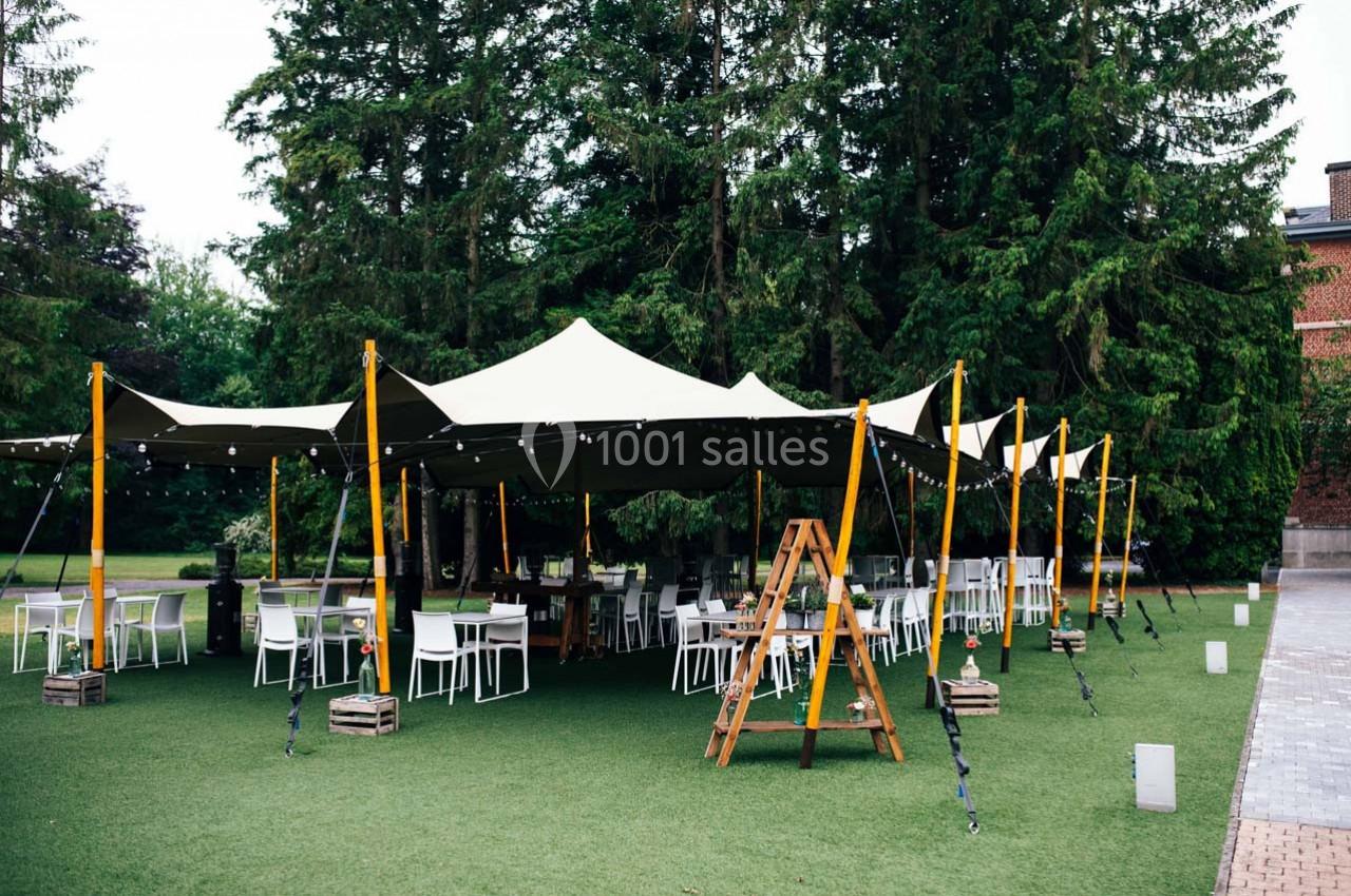 Tente beige installée sur une pelouse, entourée de chaises et tables blanches, avec des guirlandes lumineuses suspendues.