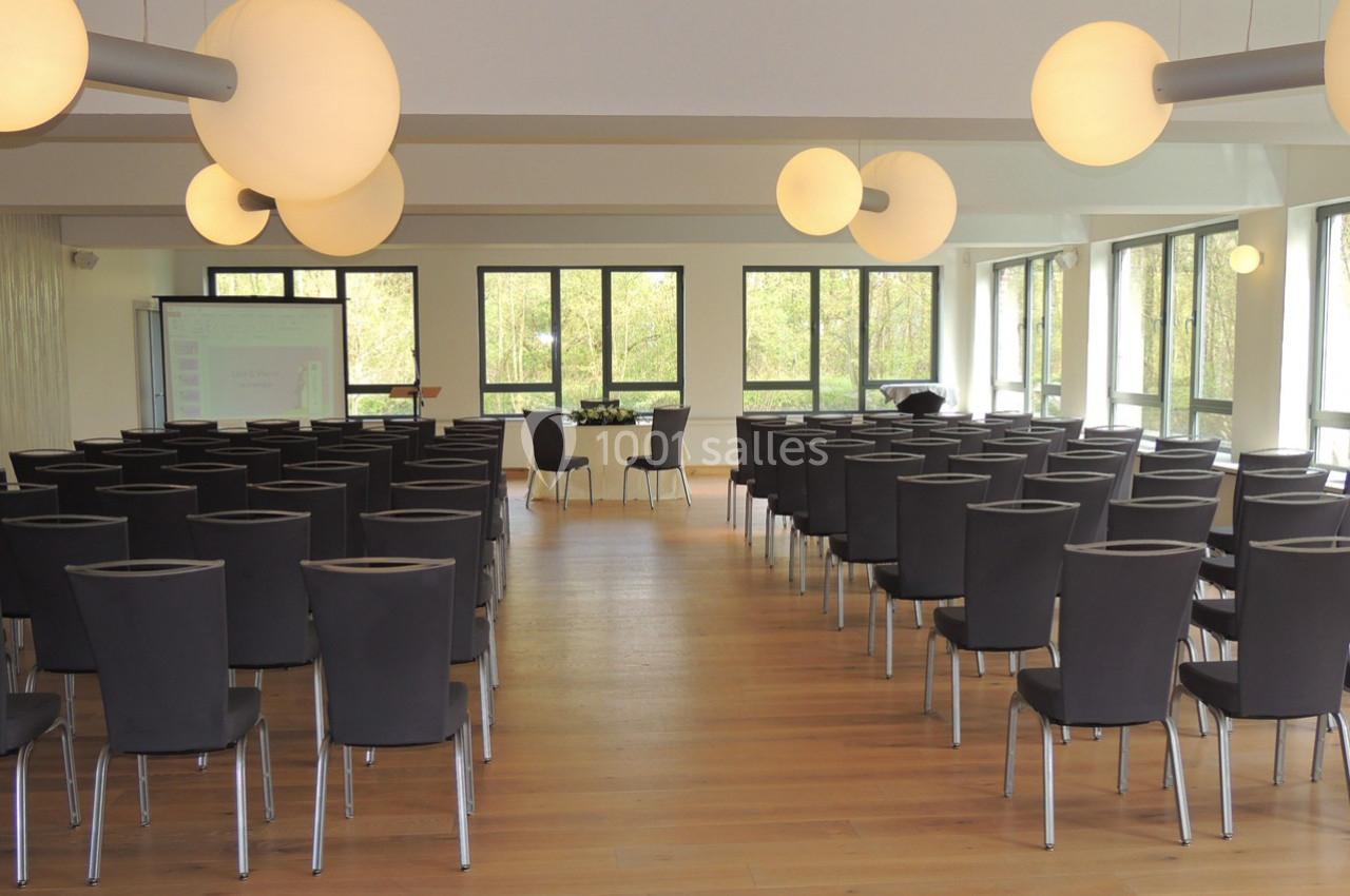 Salle de conférence lumineuse avec des rangées de chaises noires, un projecteur et de grandes fenêtres donnant sur un espace…