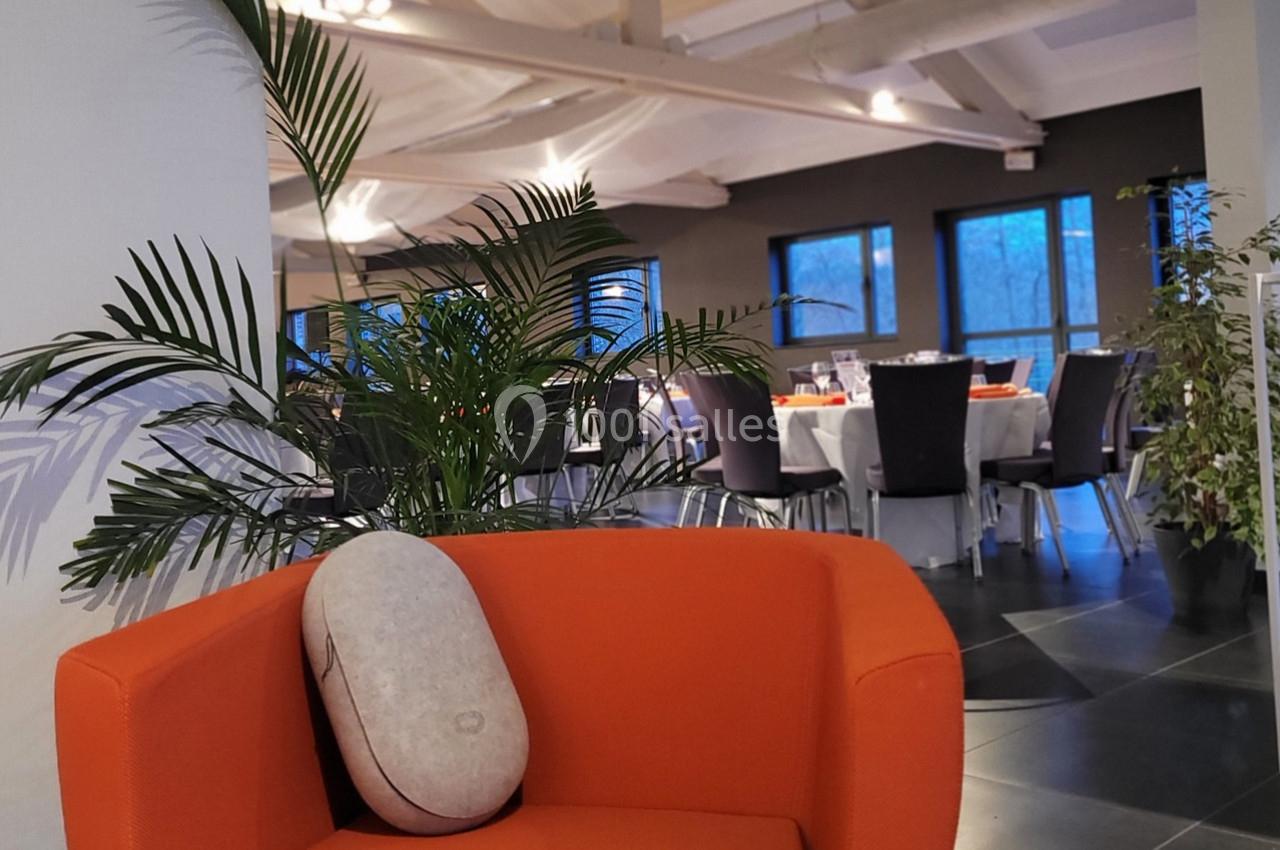 Fauteuil orange avec coussin devant une salle de réception lumineuse, tables dressées et plantes décoratives.