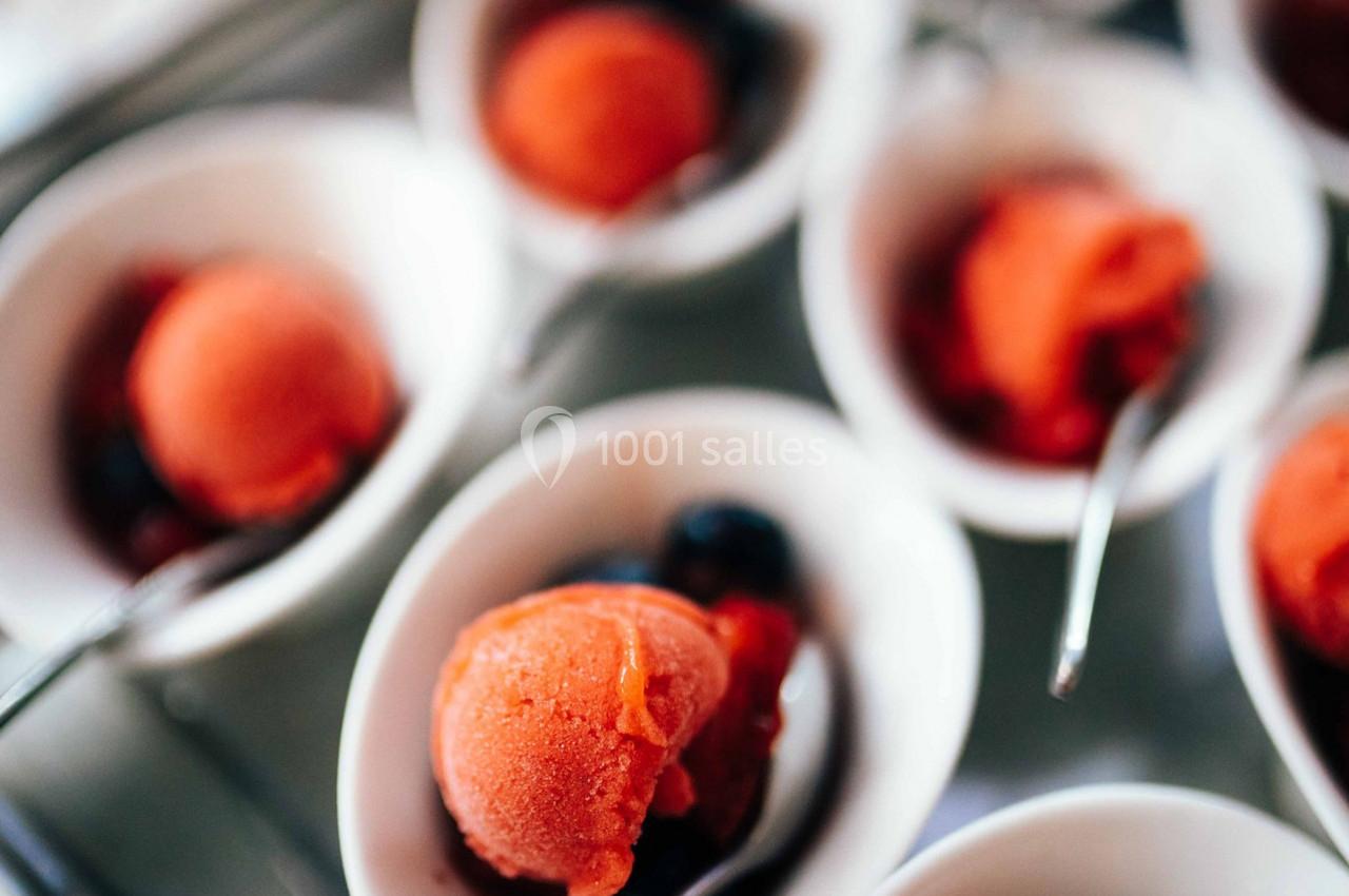 Boule de sorbet orange servie dans un petit bol blanc avec des fruits et une cuillère en métal.
