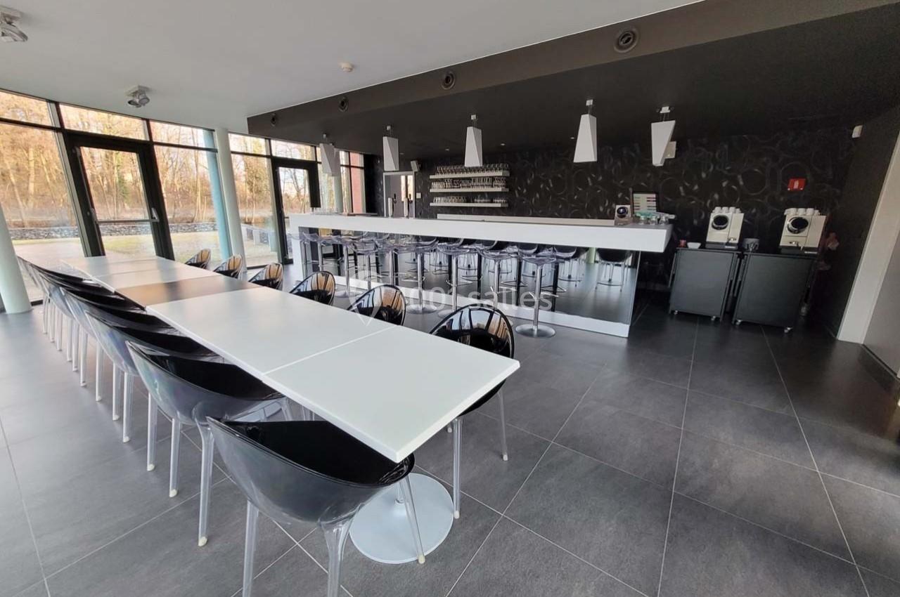 Salle moderne avec tables blanches alignées, chaises transparentes et un comptoir équipé de machines à café.