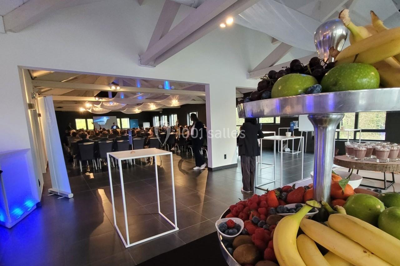 Salle de conférence lumineuse avec participants assis, vue sur un buffet de fruits frais au premier plan.