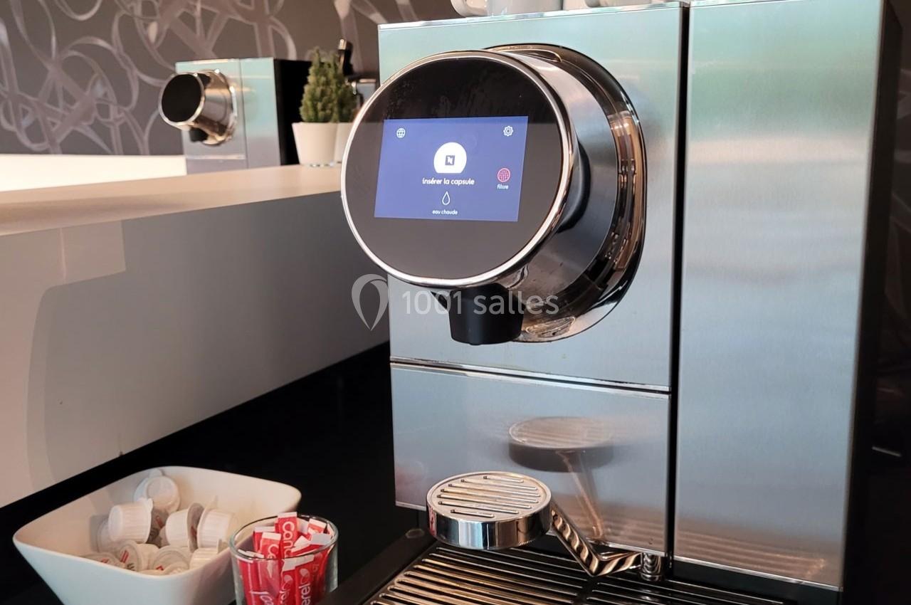 Machine à café moderne en acier inoxydable avec écran tactile, posée sur un comptoir près de dosettes et tasses.