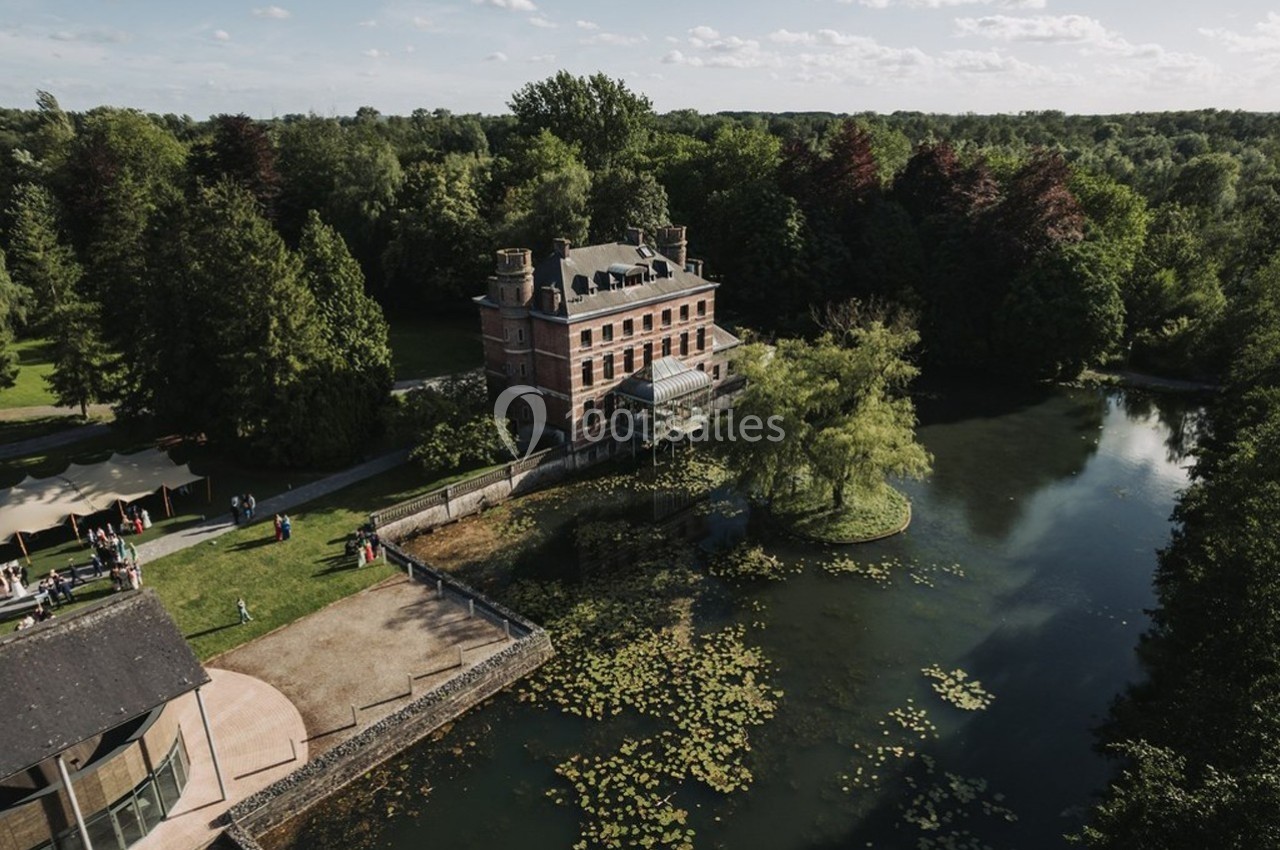 Vue aérienne d'un manoir entouré d'arbres, bordé par un étang avec des nénuphars et une pelouse aménagée.
