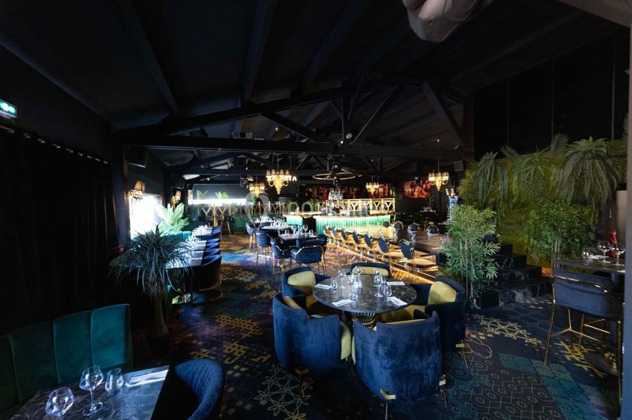 Salle de restaurant élégante avec un bar éclairé, des tables dressées, des fauteuils colorés et une décoration végétale.