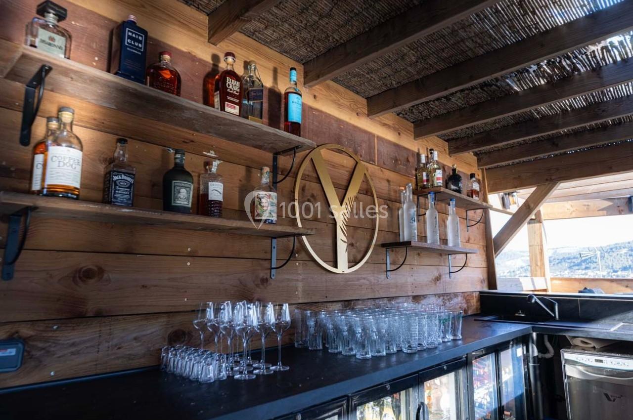 Étagères en bois garnies de bouteilles d'alcool et de verres, dans un bar à l'ambiance rustique avec vue extérieure.
