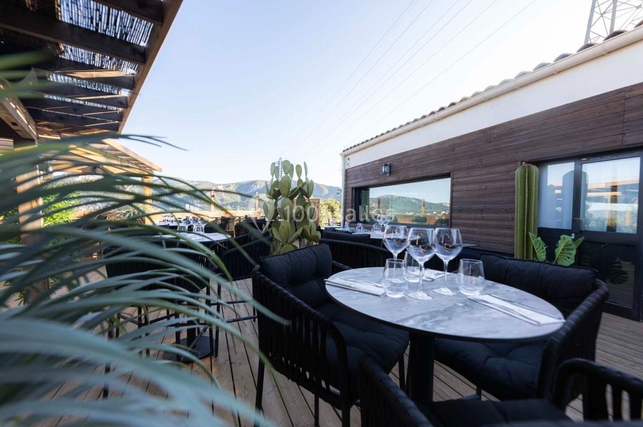 Terrasse en bois avec tables dressées, chaises noires, plantes et vue sur des collines sous un ciel dégagé.