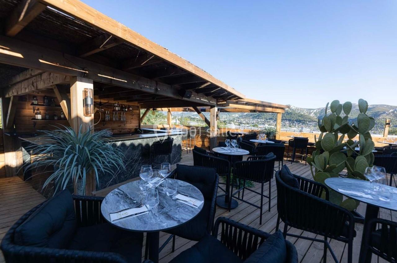 Terrasse en bois avec tables dressées, chaises noires, bar en arrière-plan et vue sur des collines sous un ciel dégagé.