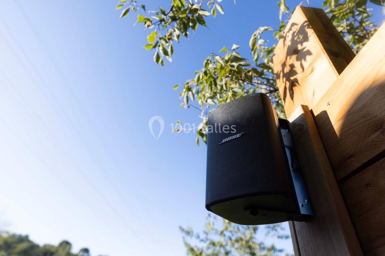 Enceinte noire fixée sur un poteau en bois, entourée de branches et sous un ciel bleu dégagé.