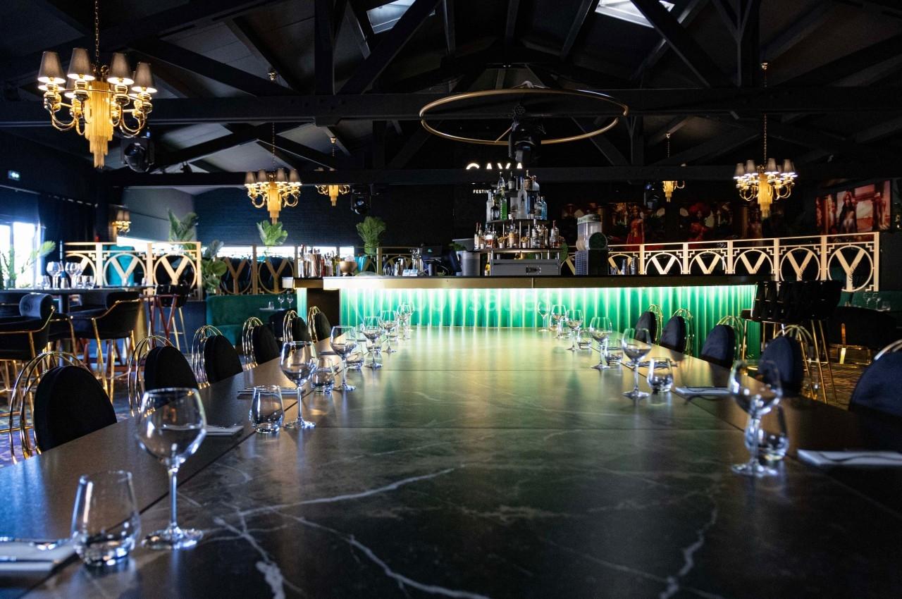 Salle de restaurant élégante avec une longue table en marbre, des chaises noires et un bar éclairé en arrière-plan.