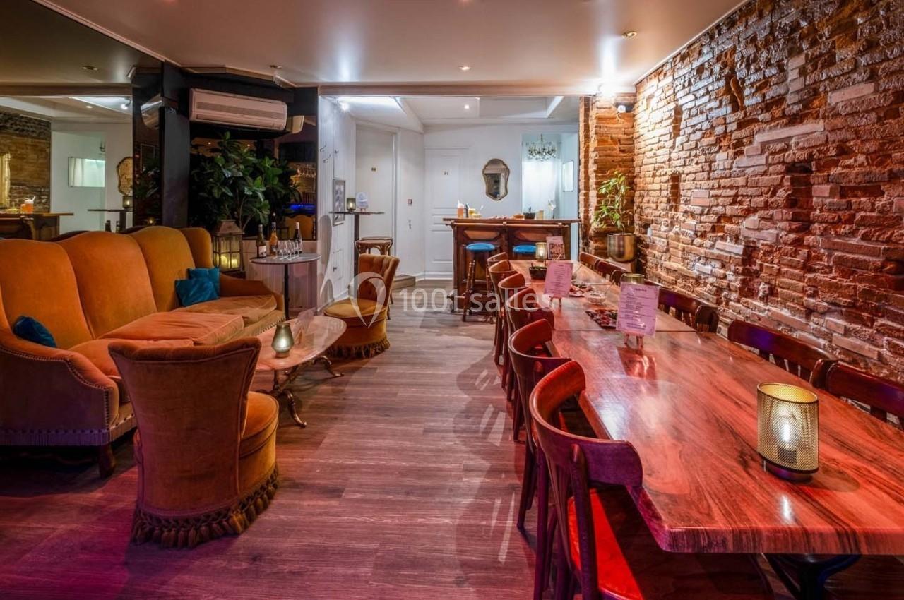 Salle chaleureuse avec murs en briques, canapé, fauteuils, grande table en bois et éclairage tamisé.